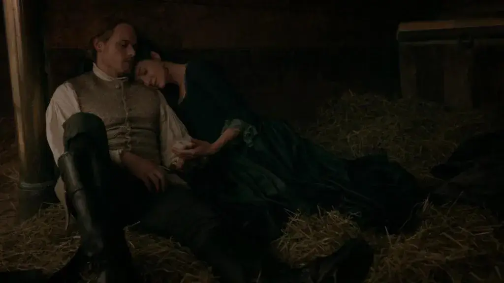 Jamie et Claire, épuisés, se reposent dans une étable, blottis l’un contre l’autre, illustrant leur tendresse et résilience après une dispute. - Saison 5

